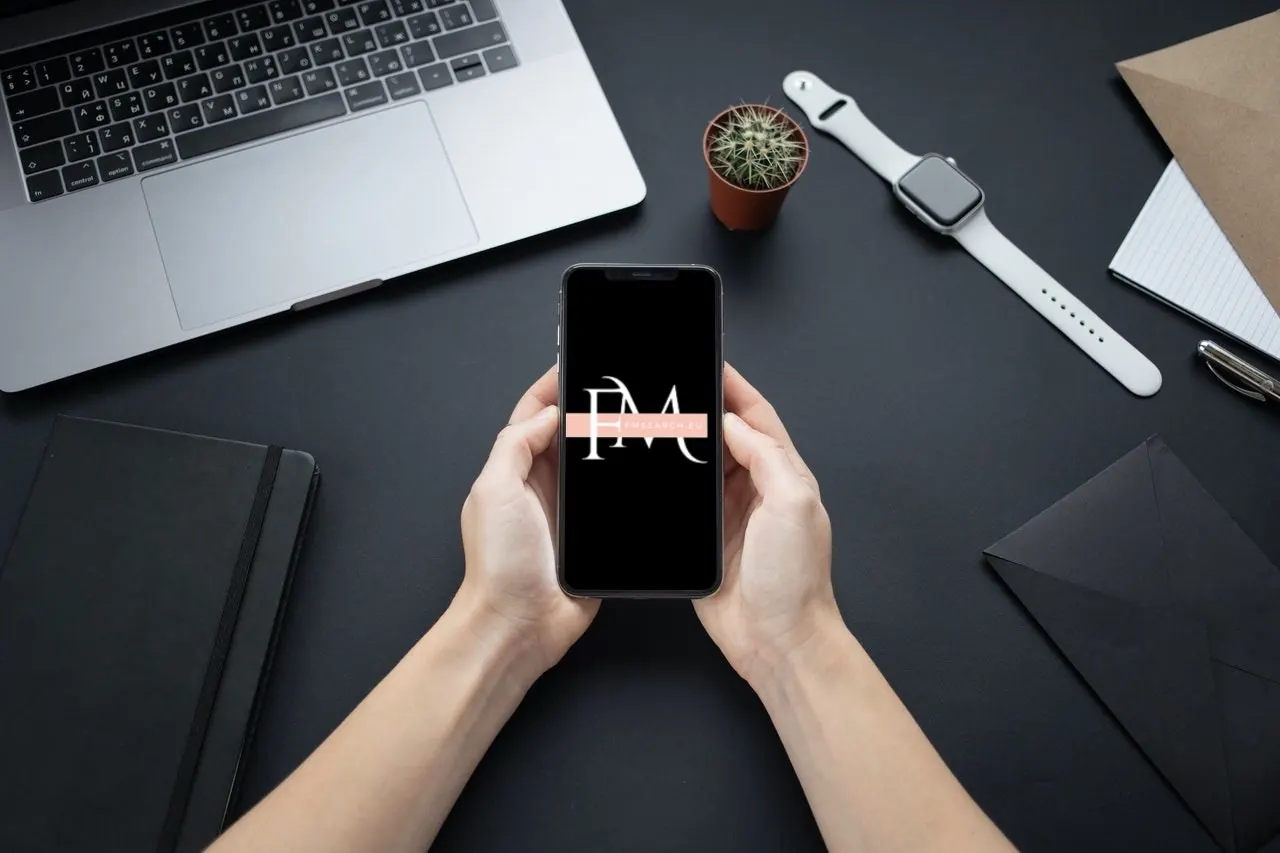 Hands holding a phone showing the FM Search logo above a laptop